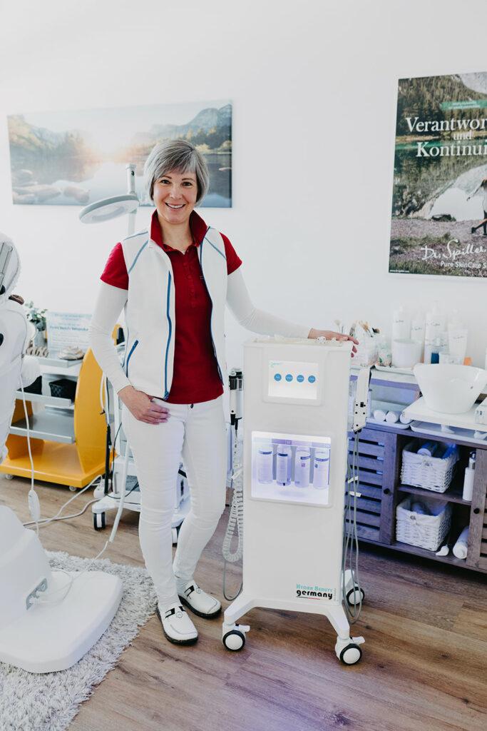 Eine Kosmetikerin steht lächelnd neben einem professionellen Hautpflegegerät in einem modernen Kosmetikstudio